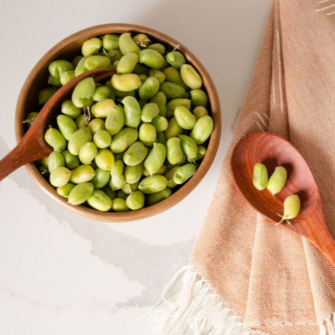 An overhead view of the wooden serving utensils in use. One spoon is dipped into a wooden bowl filled with bright green garbanzo beans, while another spoon holds a few garbanzo beans. The utensils and bowl rest on a brown tea towel textile with fringe, showcasing how the warm wood tones complement fresh ingredients and natural textiles.