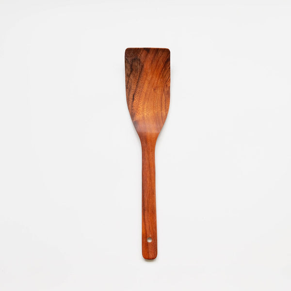 A wooden spatula or turner positioned vertically against a plain white background. The utensil is made from rich, reddish-brown wood with visible natural grain patterns. It features a wide, flat, slightly angled head for cooking and a long handle with a small hanging hole near the bottom. The polished surface shows variations in the wood tone, ranging from dark brown to amber highlights.