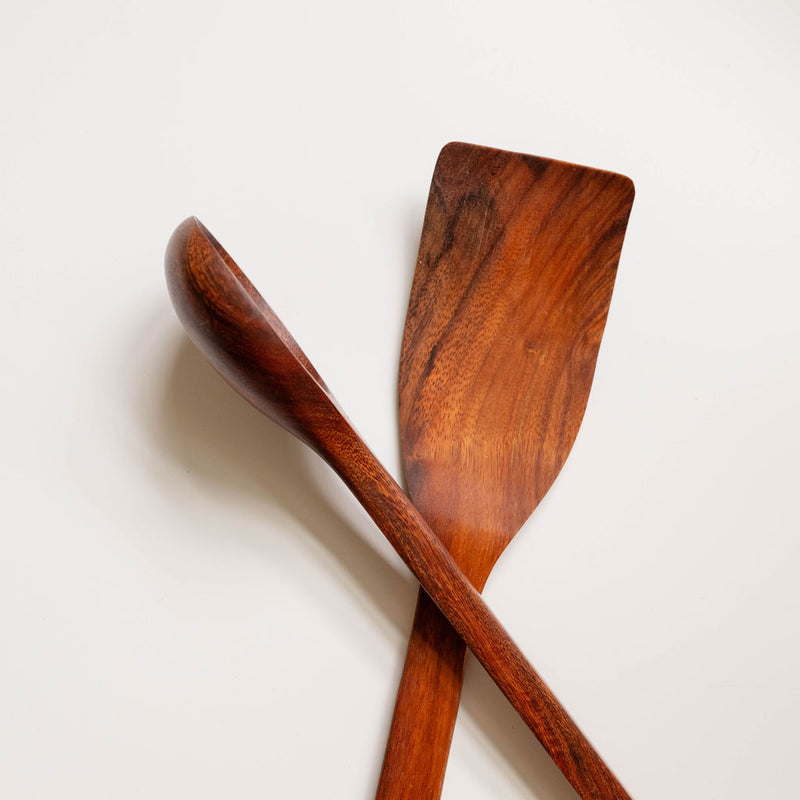 A wooden spatula  and spoon  positioned against a plain white background. The utensil is made from rich, reddish-brown wood with visible natural grain patterns. It features a wide, flat, slightly angled head for cooking and a long handle. The polished surface shows variations in the wood tone, ranging from dark brown to amber highlights.