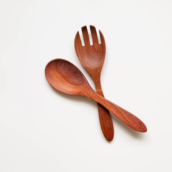 wooden serving set arranged in an X pattern against a white background. The spoon and fork are positioned to cross each other, showcasing their elegant silhouettes and the rich, reddish-brown coloration of the Mexican Rosewood with its distinctive grain patterns.