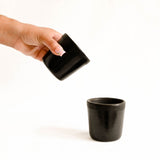 Hand pouring from a Black ceramic cup with a textured design to another cup on a white background