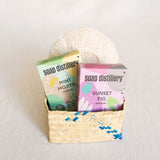 Soap Distillery's Mint Mojito Soap Bar, Soap Distillery's Sunset Fig Soap Soap Bar and VOLVERde's agave plastic-free body sponge in a handcrafted palm gift basket. 