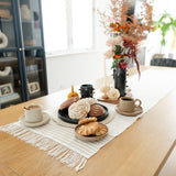 Handwoven cotton table runner in soft, neutral tones with subtle geometric patterns and fringed edges, styled on a table with conchas, plates, coffee, and flowers. 