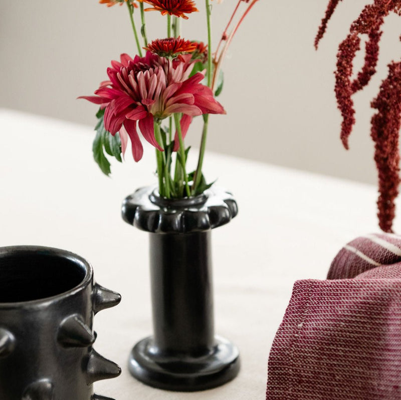 A handcrafted black clay holder with a scalloped top, featuring a smooth, metallic finish. It has a cylindrical body and a sturdy circular base, designed for dual use as a dried flower vase or candle holder. Pictured with flowers.