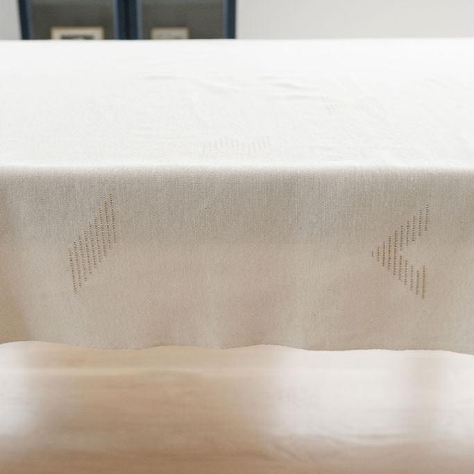 Side view of Teia Handwoven Cotton Tablecloth on a Table. Tablecloth in a beige neutral color featuring intricate geometric designs, crafted by artisans in Oaxaca. Perfect for adding a touch of elegance and tradition to your dining space. Sustainable and made from 100% Mexican cotton. Ideal for modern and rustic decor styles.