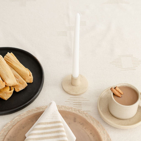 Dinner setting of an elegant Vainilla Creme Ceramic Candle Holder with a smooth, creamy finish and minimalist design. The holder features a gently curved shape, perfect for holding tapered candles, adding a touch of warmth and sophistication to any space. Next to a ceramic mug and a plate of tamales. All on a tablecloth.