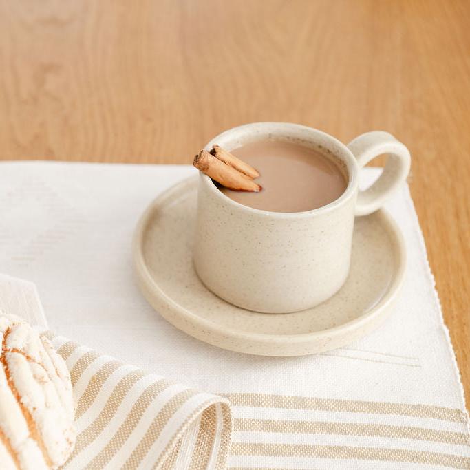 Vainilla Creme Ceramic Mug filled with coffee and cinnamon sticks with a soft, off-white finish and a smooth, matte texture. The mug features a simple, elegant design with a comfortable handle, perfect for enjoying a cozy cup of coffee or tea. The mug sits on a saucer on a placemat. 