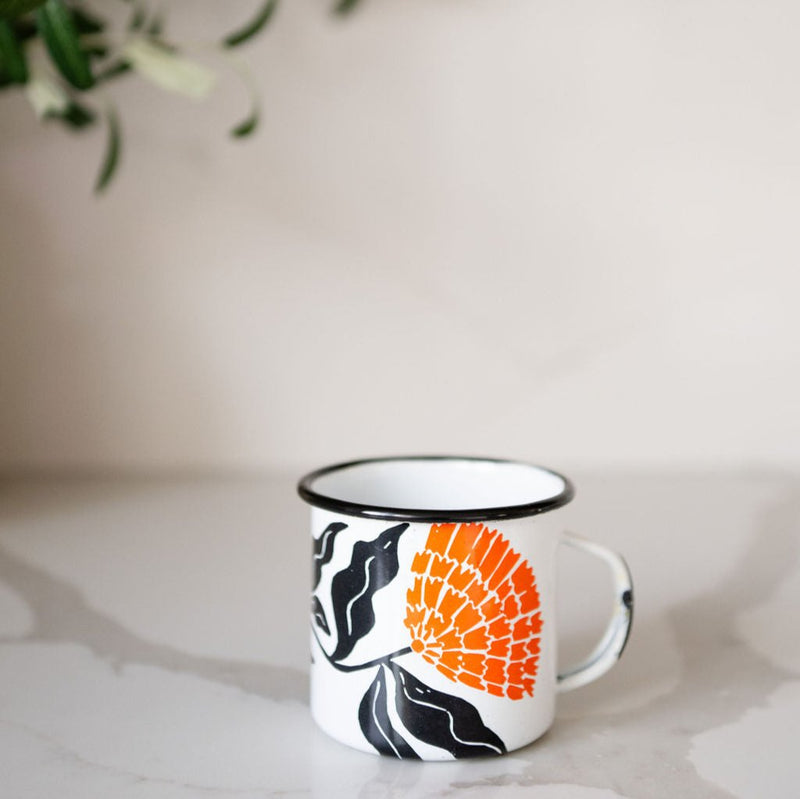 White enamel (Peltre) Cup with vibrant orange marigold (cempasúchil) designs and black leaf accents sitting on a marble countertop with a plant in the background.