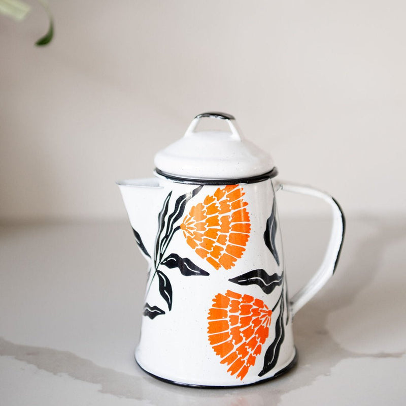 White enamel (peltre) kettle with vibrant orange marigold designs and black leaf accents, set against a marble background.