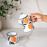 Hand holding a white enamel kettle with vibrant orange marigold designs and black leaf accents with a white enamel cup with marigold designs, set against a marbel background with a plant in the back.