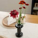 A handcrafted black clay holder with a scalloped top, featuring a smooth, metallic finish. It has a cylindrical body and a sturdy circular base, designed for dual use as a dried flower vase or candle holder. Pictured with flowers.