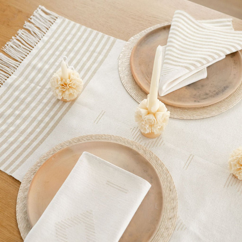 Handwoven cotton table runner in soft, neutral tones with subtle geometric patterns and fringed edges, styledon a table with plates, napkins, and candles.