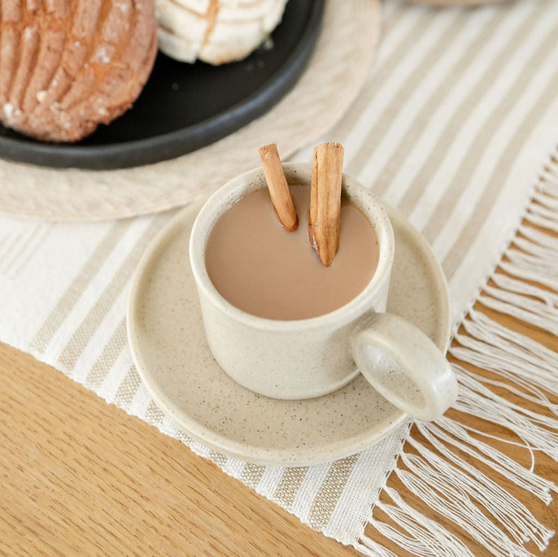 Vainilla Creme Ceramic Mug filled with coffee and cinnamon sticks with a soft, off-white finish and a smooth, matte texture. The mug features a simple, elegant design with a comfortable handle, perfect for enjoying a cozy cup of coffee or tea. The mug sits on a saucer on a placemat. 