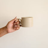 Vainilla Creme Ceramic Mug held by the handle with a soft, off-white finish and a smooth, matte texture. The mug features a simple, elegant design with a comfortable handle, perfect for enjoying a cozy cup of coffee or tea. The neutral tone complements any kitchen or table setting.