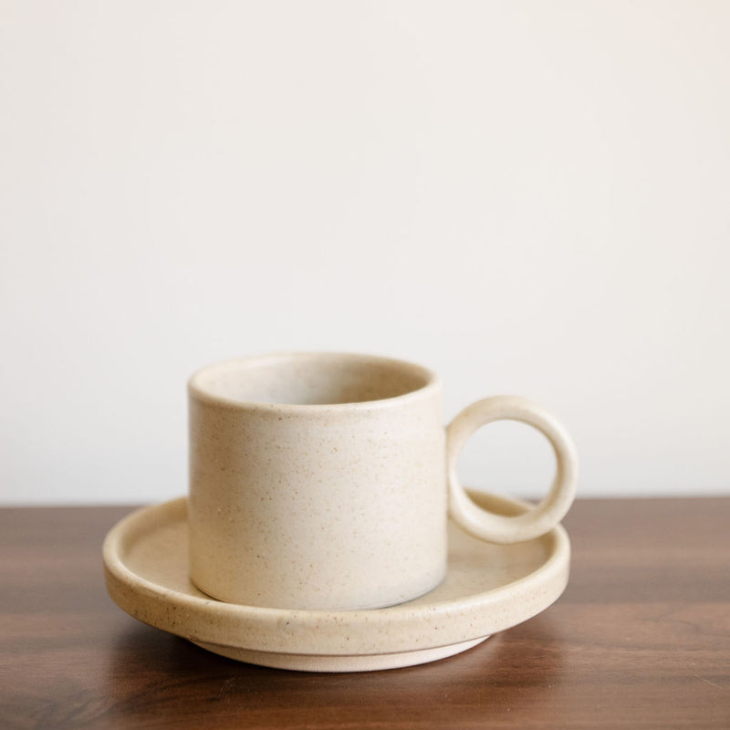 Vainilla Creme Ceramic Cup and Saucer Set featuring a smooth, cream-colored finish with subtle speckles. The cup has a rounded handle, paired with a matching saucer, both with gently curved edges for a refined look. Perfect for serving coffee or tea, this set combines simplicity and elegance.