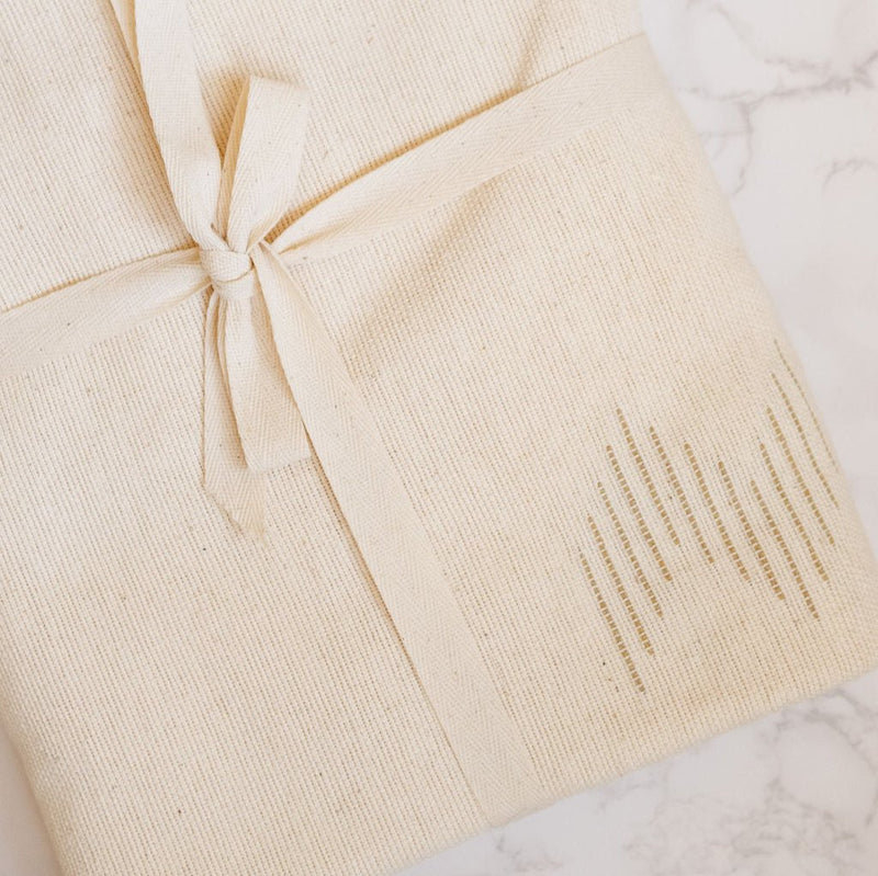 Handwoven cotton table runner in soft, neutral tones with subtle geometric patterns and fringed edges, styled folded wrapped in a bow