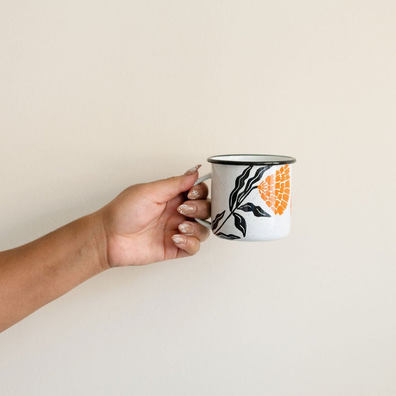 Hand holding a white enamel (Peltre) Cup with vibrant orange marigold (cempasúchil) designs and black leaf accents, set against a neutral background.