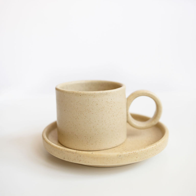 Vainilla Creme Ceramic Cup and Saucer Set featuring a smooth, cream-colored finish with subtle speckles. The cup has a rounded handle, paired with a matching saucer, both with gently curved edges for a refined look. Perfect for serving coffee or tea, this set combines simplicity and elegance.