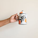 Hand holding a white enamel kettle with vibrant orange marigold designs and black leaf accents, set against a neutral background.