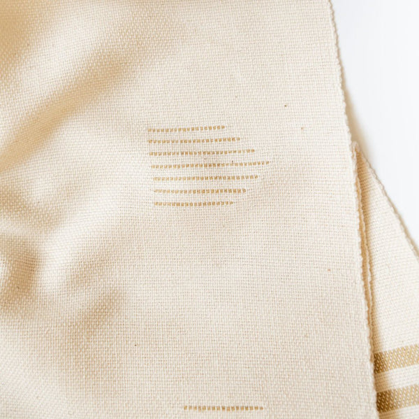 Handwoven cotton table runner in soft, neutral tones with subtle geometric patterns and fringed edges, styled against a light background