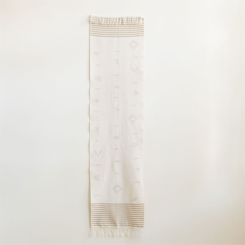 Handwoven cotton table runner in soft, neutral tones with subtle geometric patterns and fringed edges, styled against a light background