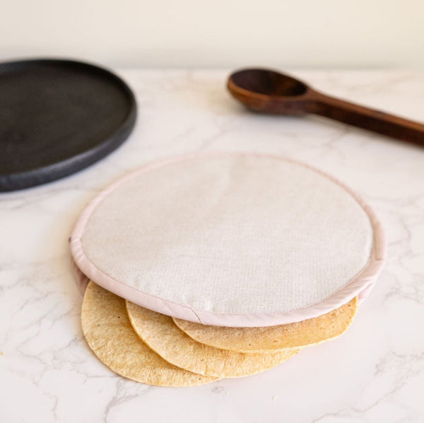 A Desert Beige Handwoven Cotton Tortilla Warmer sits elegantly on a table, showcasing its natural, earthy tones and intricate handwoven texture. Made from 100% cotton, this eco-friendly warmer adds a rustic, artisan touch to any meal while keeping tortillas warm and fresh. With a plate and spoon in the background.