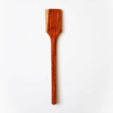 A wooden spatula or turner positioned vertically against a plain white background. The utensil is made from rich, reddish-brown wood with visible natural grain patterns. It features a wide, flat, slightly angled head for cooking and a long handle with a small hanging hole near the bottom. The polished surface shows variations in the wood tone, ranging from dark brown to amber highlights.