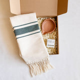 Striped cotton hand towel with fringes and a ceramic  soap dish with a organic soap bar in an open gift box box on a white surface
