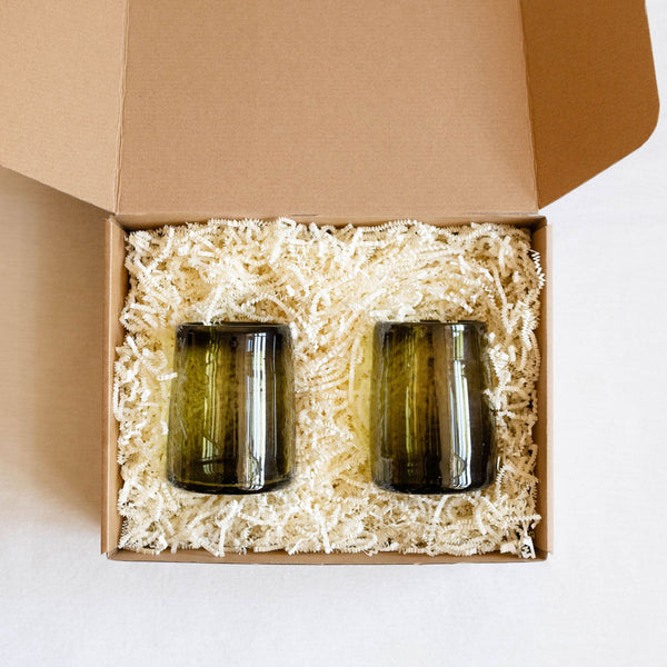 Two recycled green glass tumblers in a kraft gift box with shredded paper.