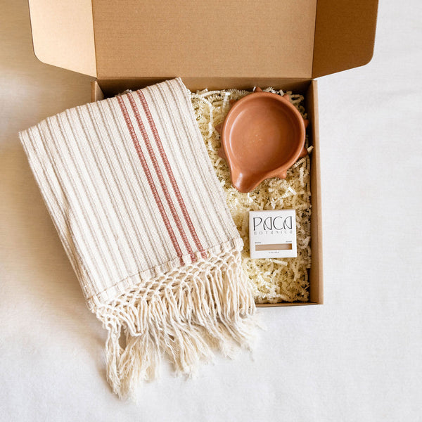 Gift box containing a handwoven cotton towel with cinnamon-colored stripes, an Oaxacan terracotta spiked soap dish, and a Marigold turmeric and calendula soap bar arranged on natural crinkle paper.