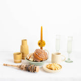 A styled tabletop arrangement on a white surface against a white background, featuring a light green cake pedestal with assorted pastries, a tall orange candle with yellow flower accents, two clear glass champagne flutes, a cream mug, a small beige vase, a wooden molinillo, and a small plate with a pastry