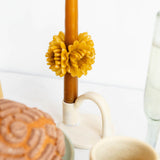 A close-up lifestyle shot of a candle holder featuring a brown candle wrapped in a yellow flower-shaped wax sleeve, set on a white surface next to a beige ribbed mug and a pastry.