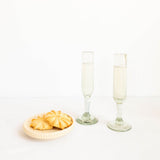 Two clear glass champagne flutes filled with a pale golden beverage, standing on a white surface beside a small beige plate holding two golden cookies, against a clean white background.