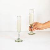 Two clear glass champagne flutes on a white surface against a white background, with a hand holding one flute.