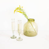 An olive green glass vase with a single yellow flower stem, next to two clear glass champagne flutes, on a clean white background.