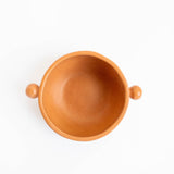 A top-down view of a small terracotta clay bowl with two small side handles, shown on a plain white background. Burnished ceramics from Nahuatl community in Michoacan, Mexico.