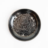 A round, black Huancito Mexican burnished ceramic hand-painted plate with floral designs, styled on a striped tablecloth with fresh vegetables.