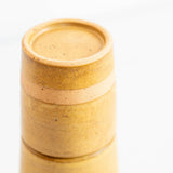 A close-up of a cylindrical ceramic bedside carafe with a textured, speckled matte finish in warm sandy beige and light brown tones, set against a clean white background.