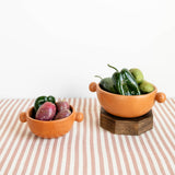 Two terracotta-colored Mexican ceramic bowls with round side handles on a striped tablecloth against a white background. Handcrafted burnished ceramic bowls from Michoacan