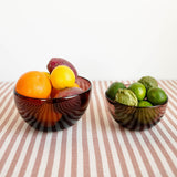 Two amethyst glass bowls filled with fruit, including oranges, lemons, limes, and a purple fruit, set on a striped tablecloth against a white background.