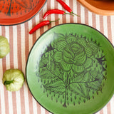 A green Huancito burnished ceramic plate with hand-painted floral designs, one red and one green, styled on a striped tablecloth with tomatoes, and chilis.