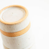 A close-up of a handmade ceramic container or vase with a textured off-white glaze and a warm tan band around the middle, set against a bright white background.