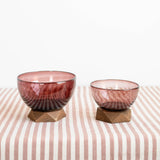 Two small amber-brown handblown glass bowls with geometric wooden bases, displayed on a tablecloth against a white background.