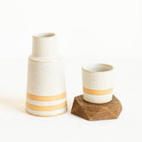 A ceramic Bedside Carafe set featuring a tall, conical vase and a matching small cup/lid, both in an off-white gray speckled glaze with a warm ochre band near the base, displayed on a clean white background. The cup sits on a geometric wooden coaster.