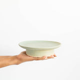 A hand holding a ceramic stand pedestal with a light sage green hue against a plain white background.