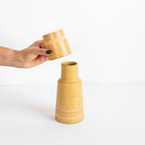 A hand with dark nail polish holds a small matching ceramic lid above a larger tapered ceramic bedside carafe featuring horizontal terracotta bands, set against a clean white background.