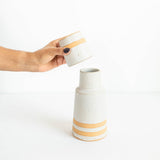 A hand with dark nail polish holds a small speckled ceramic cup above a larger matching vase, both in a warm off-white glaze with two horizontal tan stripes, against a clean white background.