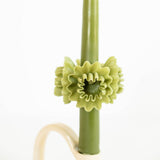 A green beeswax taper candle with a sculpted olive-green flower ring around the middle, displayed on a cream-colored stand against a plain white background.