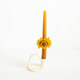 Oaxacan Beeswax taper candle in a warm golden-amber color, decorated with a sculpted yellow flower at the center and resting in a white ceramic holder against a plain white background.
