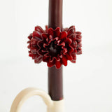 Close-up of a burgundy red taper candle with a textured, ruffled flower decoration in the center against a plain white background.